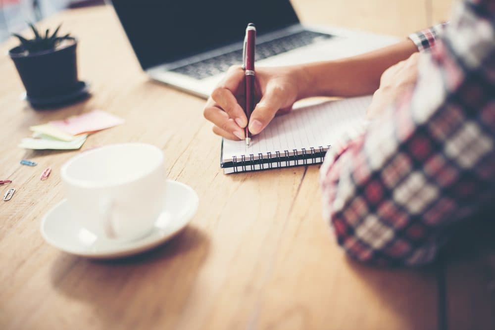 Writer taking notes in a notebook at a desk with a laptop, representing contributing articles to Reflax